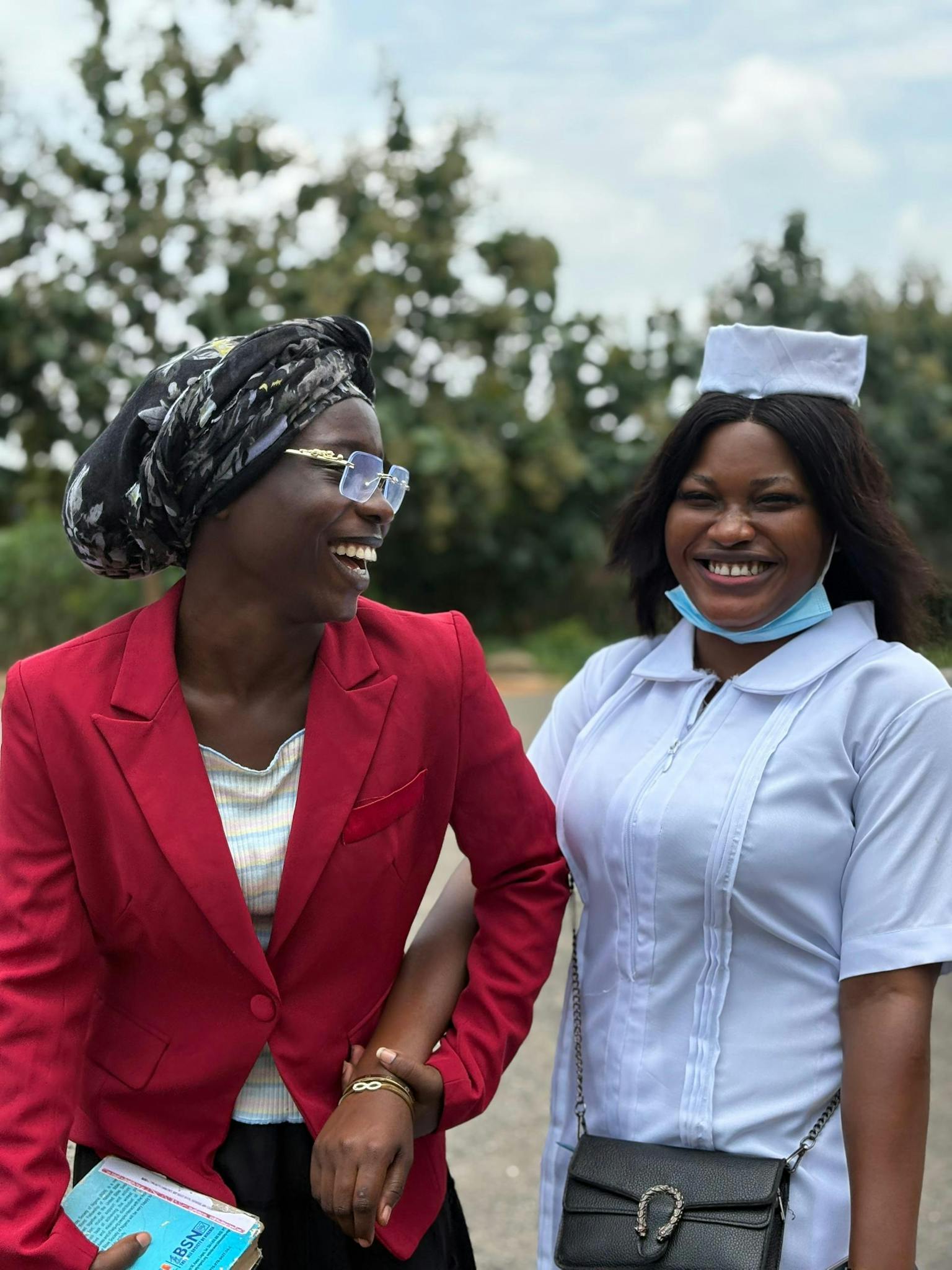 Two women laughing outdoors, one in a red blazer, the other in a nurse uniform.